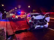 A small white vehicle with major front-end damage sits in the road with a police SUV behind