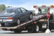 A tow truck carrying a smaller grey sedan parked next to a red stop sign, 
